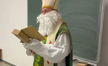 Der Nikolaus mit langem Bart & festlichem Gewand steht vor der Tafel eines Klassenzimmers und liest aus einem dicken Buch vor.